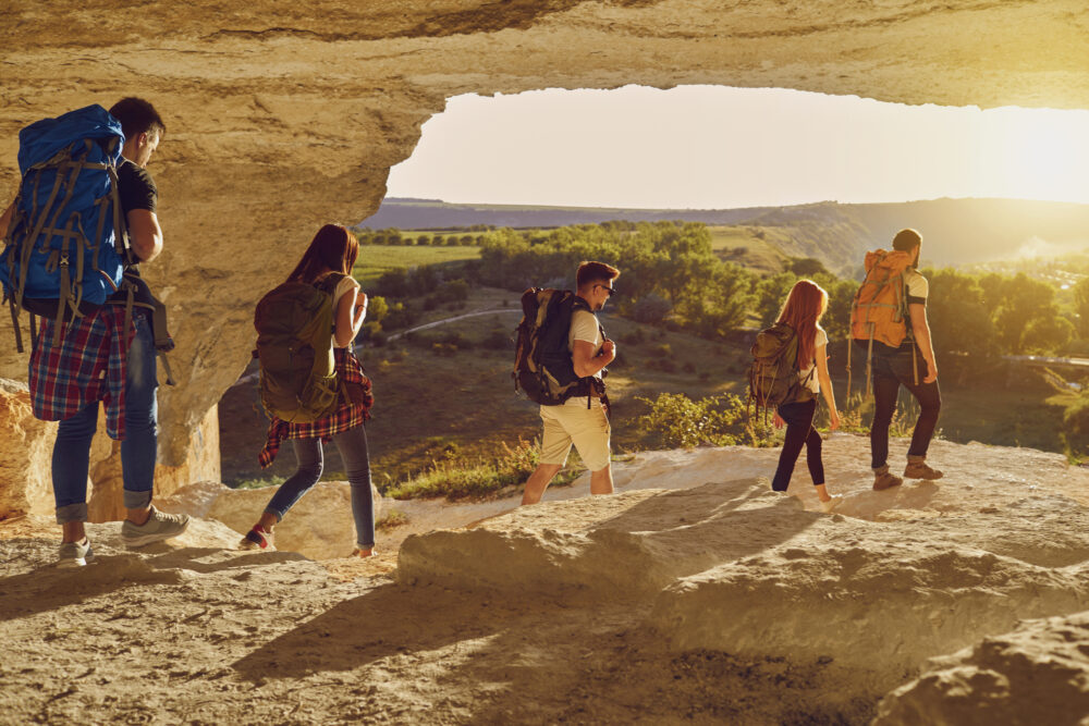 Group of young friends hikers hiking in natural rocks with backpacks in row together during summer vacations on sunny clear day, rear view. Hiking, traveling with backpack, vacations concept