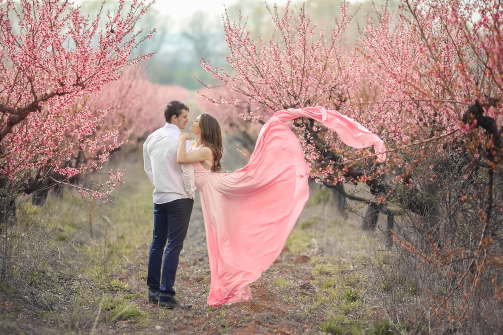 Romantic bridegroom kissing bride on forehead while standing against wall covered with pink flowers