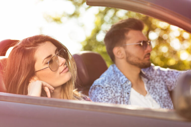 Young Couple Driving Along Country Road In Open Top Car.